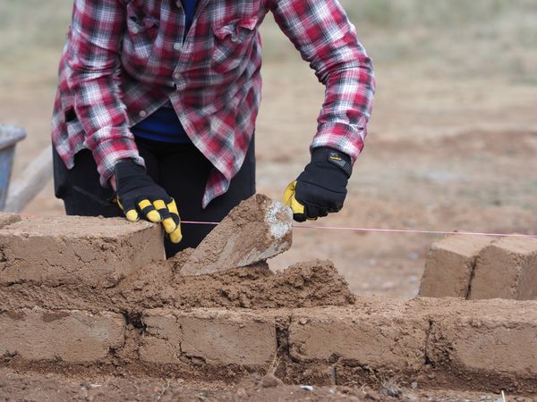 A worker trowels mud across a brick before placing another one, which will be leveled to the height of the pink string.